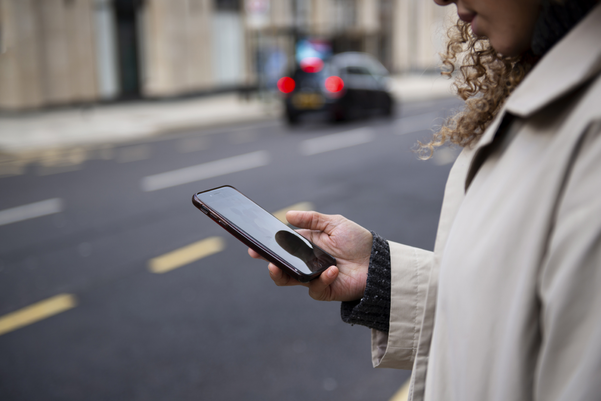 zoom su mano con smartphone di una persona su marciapiede e vista di automobile in lontananza